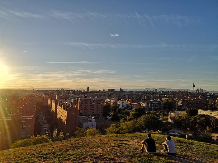 Guía para vivir en Vallecas: zonas recomendadas, transporte y calidad de vida 4 Dos personas sentadas en el césped del Parque de las Siete Tetas (Cerro del Tío Pío) contemplando el skyline de Madrid y Torrespaña bajo la luz dorada del atardecer