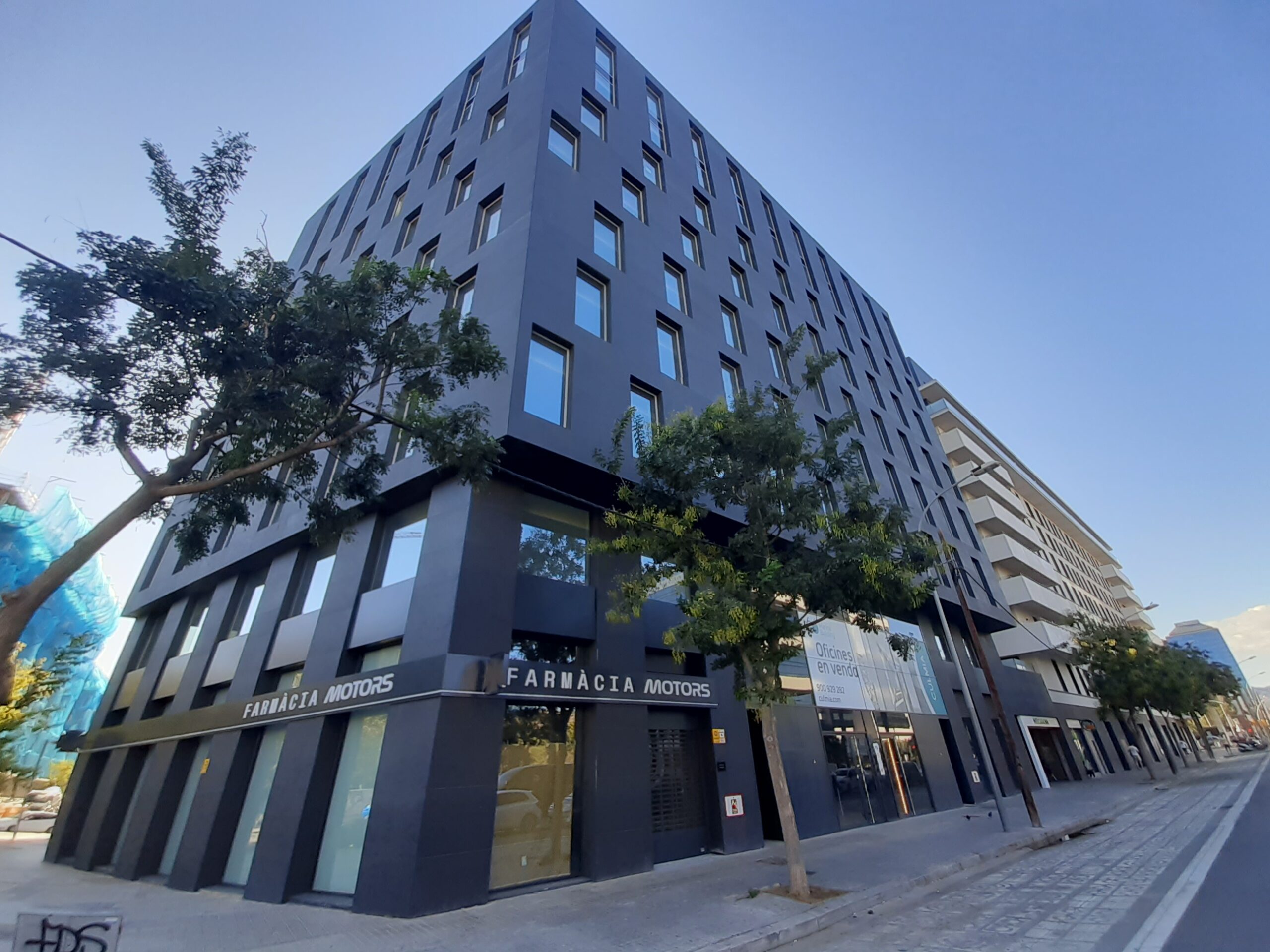 Vista exterior de un edificio de oficinas moderno de color negro con grandes ventanales y un local de Farmàcia Motors en la planta baja